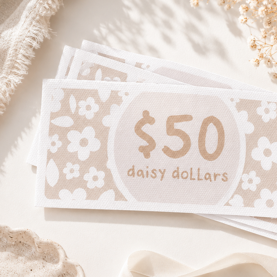 A stack of decorative $50 gift cards labeled 'daisy dollars' with a floral pattern, styled on a light surface with dried flowers, a frayed textile, and ribbon.