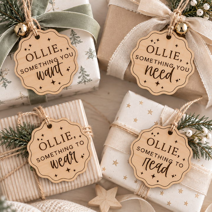 Four wrapped gift boxes arranged on a tabletop with decorative ribbons, pine branches, and small gold ornaments. Each package has a round wooden tag reading “OLLIE, SOMETHING YOU need” or “OLLIE, SOMETHING YOU want” in a warm beige holiday-style font.
