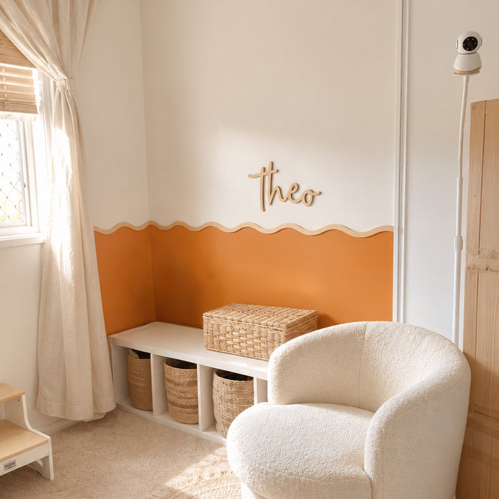 A cozy nursery corner featuring a round, cream bouclé armchair on a woven jute rug, next to a white bench with storage baskets and a wicker box. The wall is painted with a wavy orange stripe and a wooden &
