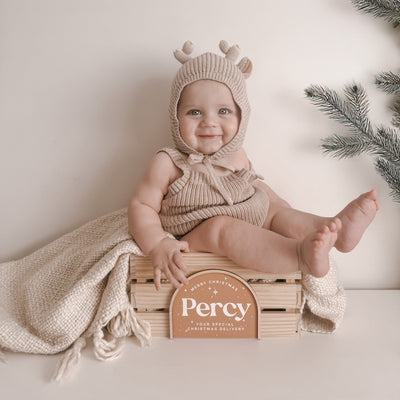 Baby in a knitted outfit sitting on a wooden crate with a sign reading 'Percy' against a neutral background.