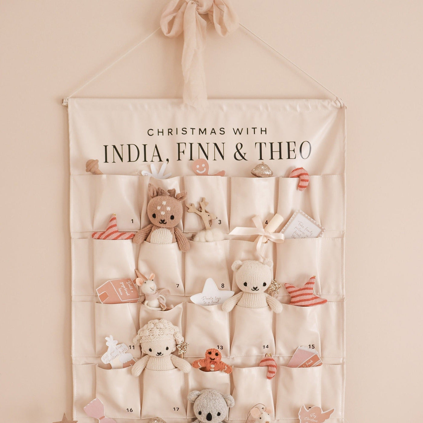A beige vegan leather Christmas advent calendar with wooden and acrylic name design, hanging on a wall with various small toy figures and decorations inside the pockets.