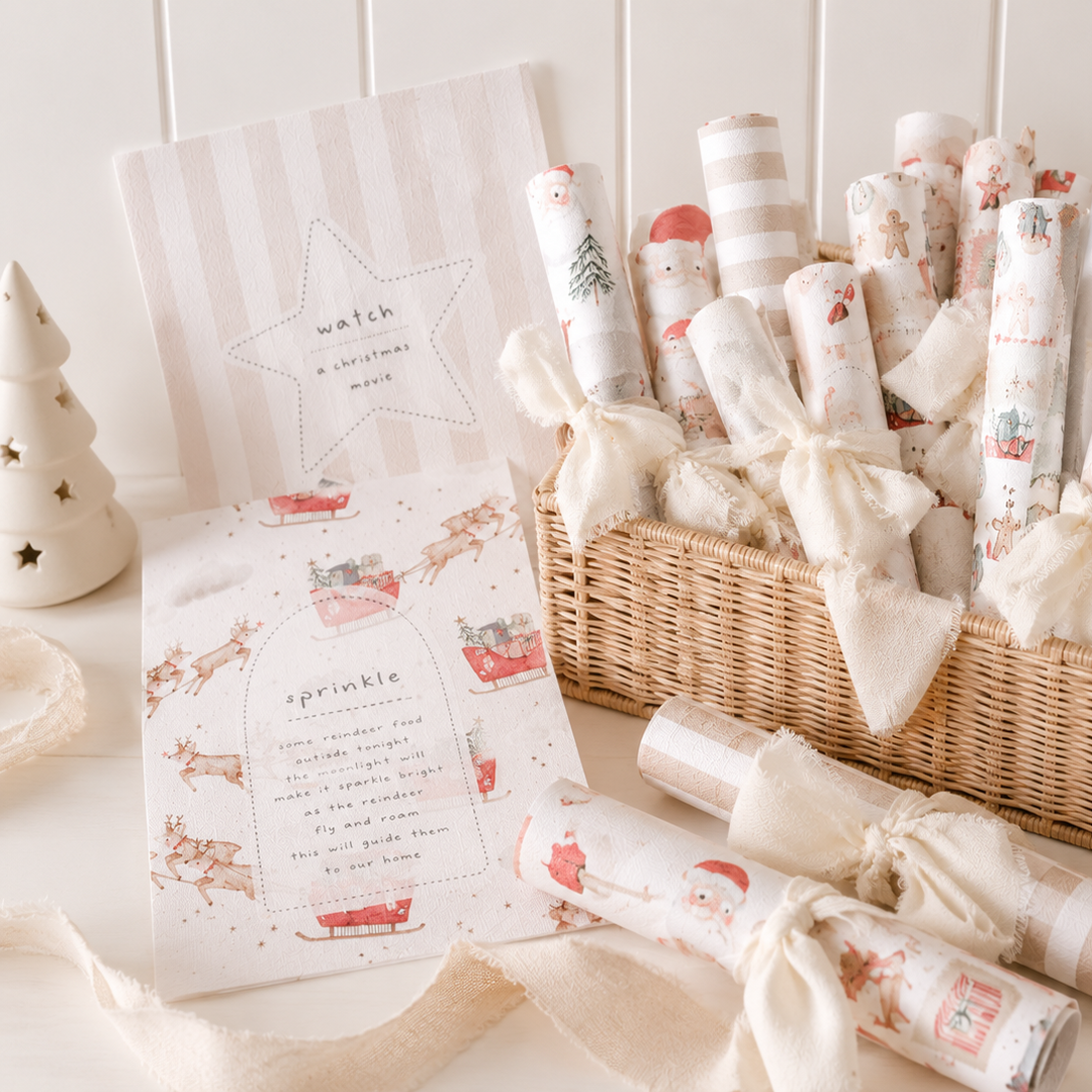 A wicker basket filled with rolled Christmas-themed wrapping paper and gift bags tied with cream ribbons, alongside matching printed cards and a small decorative tree on a light tabletop.
