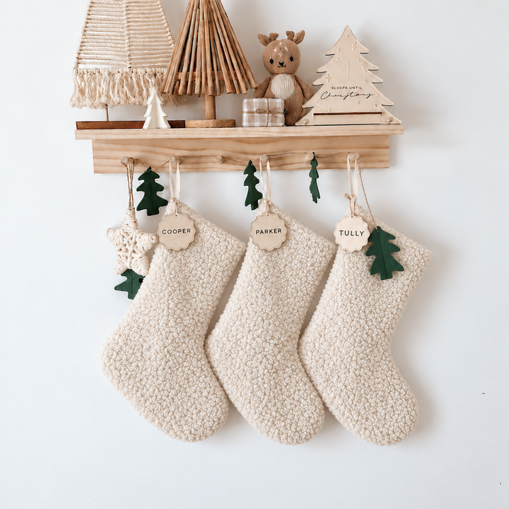 A wooden wall-mounted shelf decorated with small Christmas trees, a teddy bear, and three cream-colored textured hanging stockings with green felt trees and personalized name tags reading &