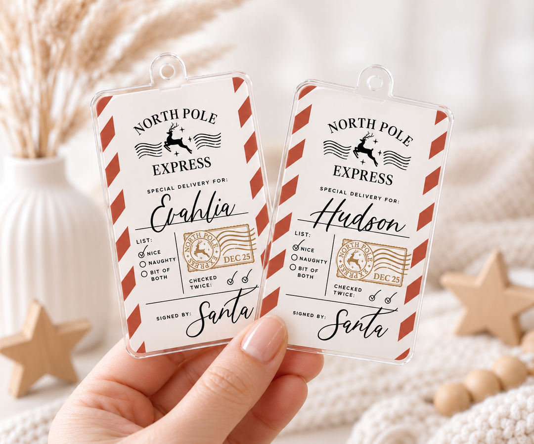 A hand holds two clear plastic holiday-themed luggage tags with a vintage airmail design. The tags feature red and white diagonal stripes, black text reading 'NORTH POLE EXPRESS', and gold-toned postage stamp graphics.